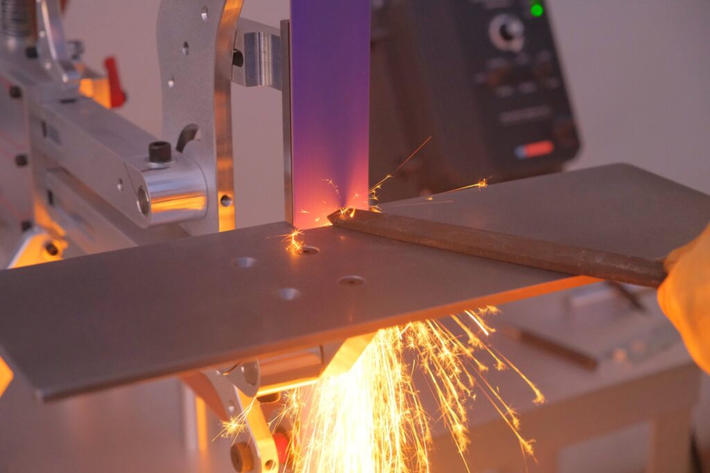 A purple sanding belt is chewing through a steel rectangle as it's pushing into the rapidly spinning sanding belt. A lot of sparks are visible.